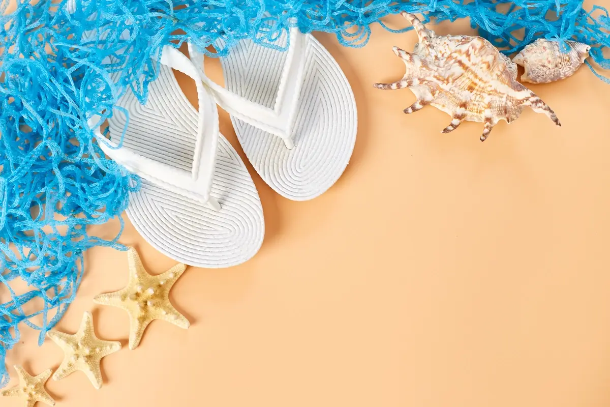 White woven flip-flops, blue net, seashell and starfish scattered on warm peach sand