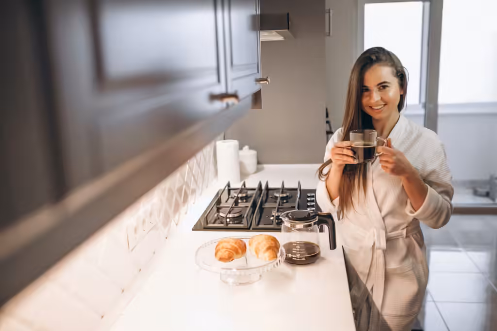 hotel voor lange reis: lachende vrouw in witte badjas met koffie en croissants aan het aanrecht