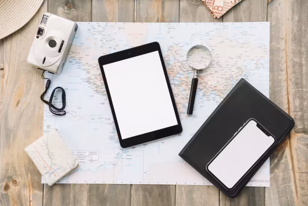 independent luxury travel: a flat lay of tablet, smartphone, notebook, compact camera and magnifying glass on a world map